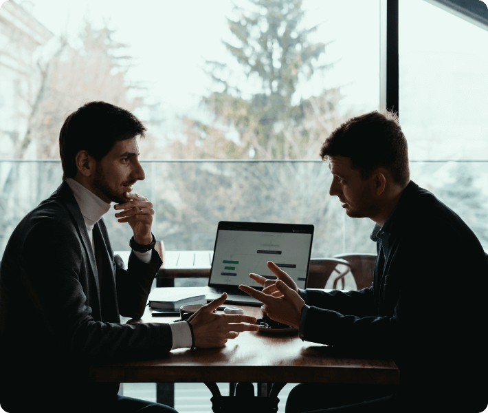 Two men at a table, collaborating on a project with a laptop open in front of them.