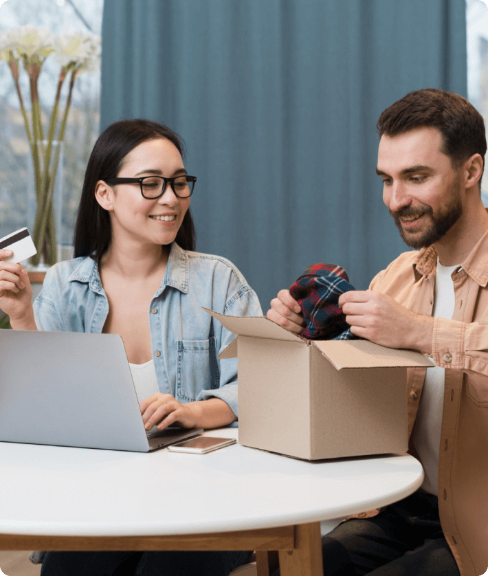 A man and woman are seated at a table, focused on a laptop, with a box placed in front of them, indicating a work session.